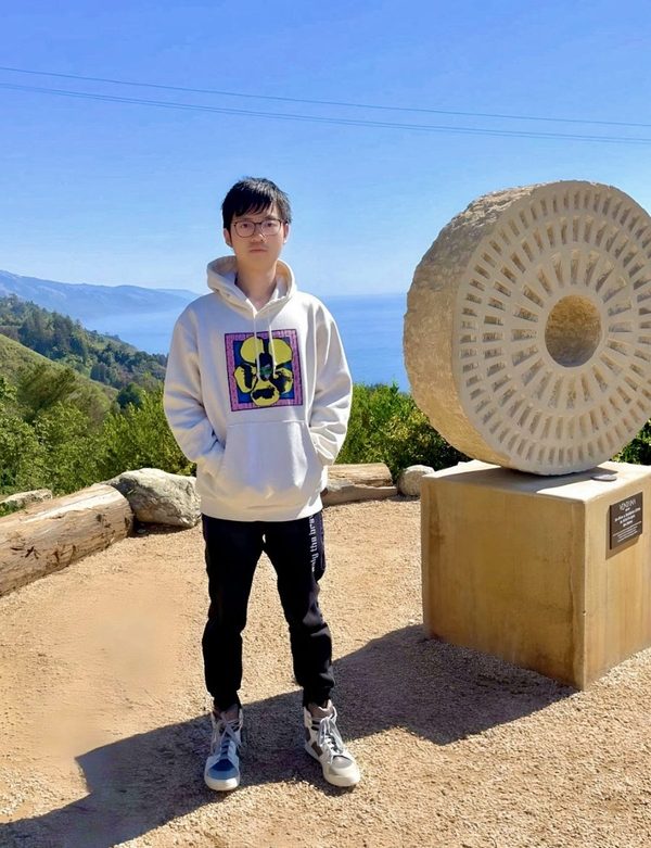 Zheng Zhu standing outdoors beside a circular stone sculpture at Big Sur, with the coastline in the background
